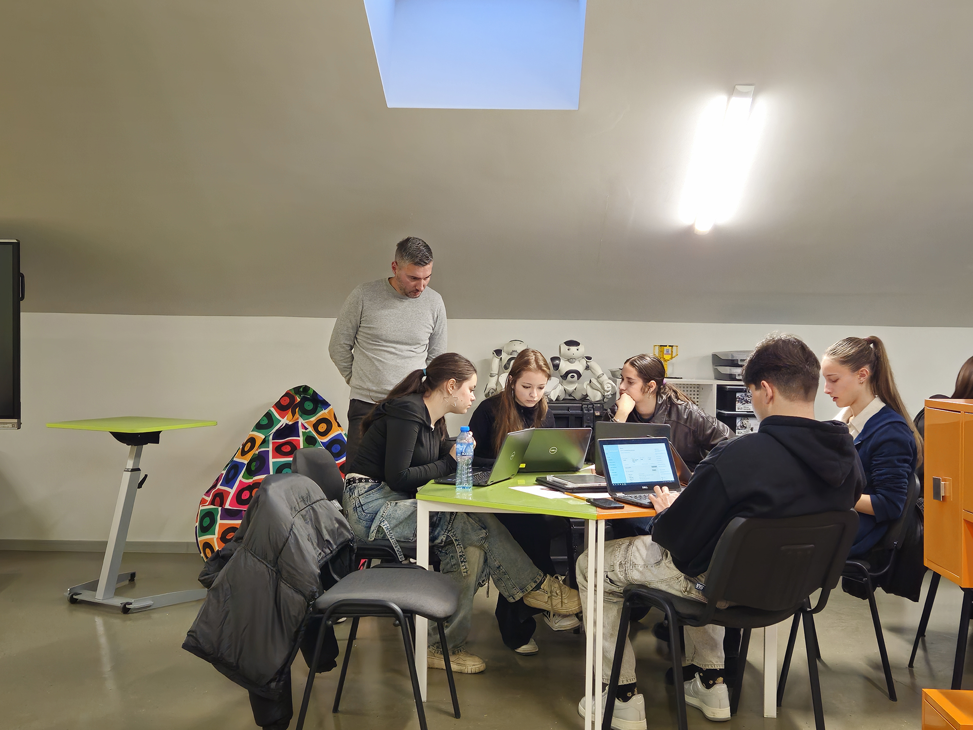 Students and instructor at triangular table with laptops, robotics in background